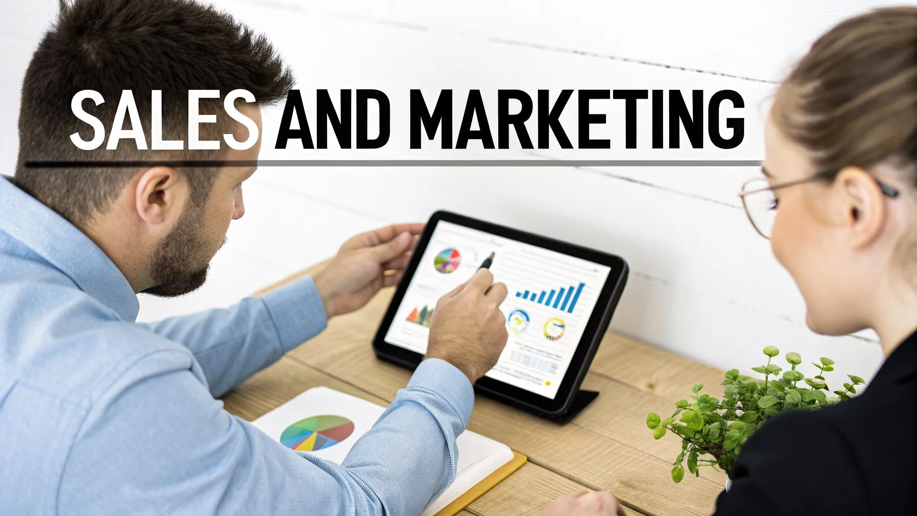 Two colleagues analyze sales and marketing data on a tablet, displaying charts and graphs.